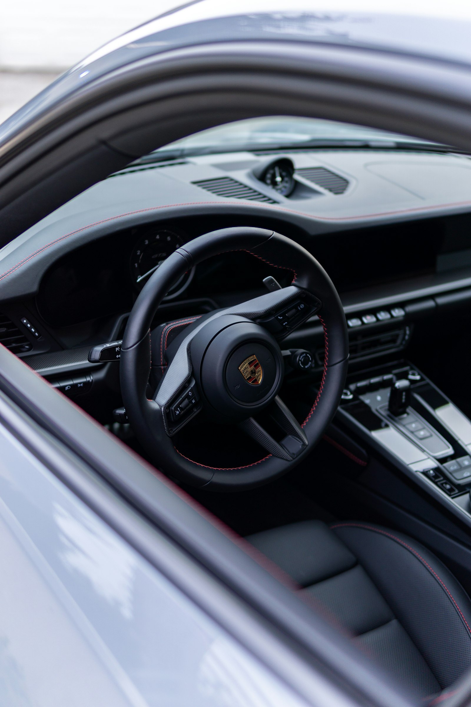 A black steering wheel with red stitching and a dark leather seat, seen through an open car window